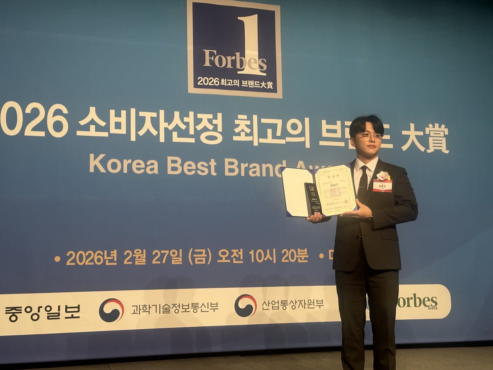포브스 코리아 2026 소비자 선정 최고의 브랜드 대상 시상식 현장. 최광수 대표가 GSLT 이름이 적힌 인증서와 상패를 들고 무대에 서 있고, 배경 화면에 '2026 소비자선정 최고의 브랜드 大賞 Korea Best Brand Awards', 2026년 2월 27일 금요일 오전 10시 20분, 중앙일보·과학기술정보통신부·산업통상자원부·Forbes Korea 로고가 보인다.